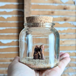 May include: A clear glass jar with a cork stopper containing a layer of light brown sand at the bottom. Inside the jar, two small brown figurines with hooded robes and glowing yellow eyes stand side by side on the sandy surface.