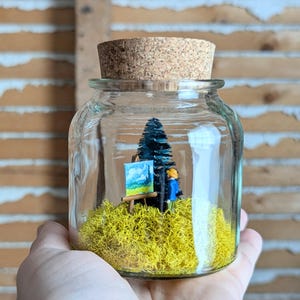 May include: A miniature art scene within a glass jar, depicting a small artist painting a landscape. The scene includes a tiny easel with a painting, a dark green tree, and yellow moss. The jar is sealed with a cork stopper.