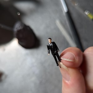 May include: A small black figurine of a person wearing a suit. The figurine is standing on a finger.