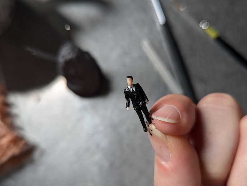 May include: A small black figurine of a person wearing a suit. The figurine is standing on a finger.