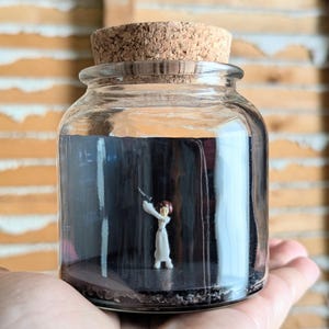 May include: A clear glass jar with a cork stopper containing a miniature figurine of a person in a white dress holding a small black blaster. The interior of the jar features a dark black background and a dark base, creating a high-contrast display.