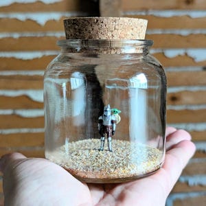 May include: A clear glass jar with a cork stopper containing a miniature silver-coloured armoured figure with a small green creature on its shoulder, standing on a layer of light brown sand inside the jar, creating a small display piece.