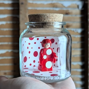 May include: A glass jar with a cork stopper, housing a red Lego figure with white polka dots. The figure is set against a backdrop of white paper with red polka dots. The jar rests on small white pebbles. The jar is clear.