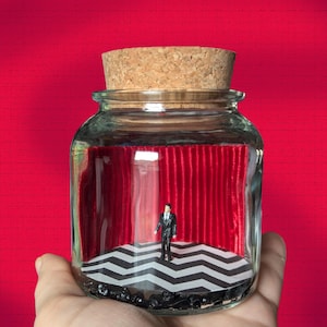 May include: A glass jar with a cork lid containing a miniature diorama of a man standing in front of a red velvet curtain and a black and white checkered floor.