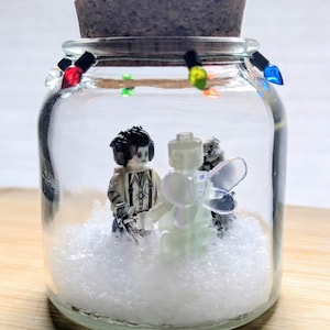 May include: A clear glass jar with a cork lid containing two miniature figures standing in artificial snow. One figure has dark hair and a white outfit, while the other is translucent with wings. A string of small multicoloured lights encircles the jar.