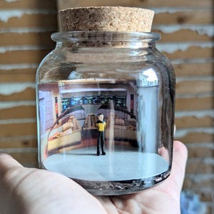 May include: A clear glass jar with a cork stopper containing a miniature figurine of a character in a yellow and black uniform standing on a white base in front of a printed backdrop depicting a futuristic spaceship bridge interior scene.
