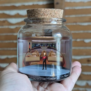 May include: A glass jar with a cork stopper, featuring a miniature Star Trek diorama. Inside, a figure stands on a blue platform, surrounded by a detailed bridge interior. The jar is held in a hand.
