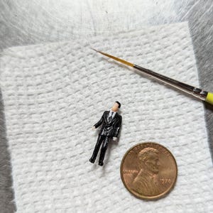 May include: A small, black figurine of a person wearing a suit standing on a white paper towel next to a 1976 US penny.