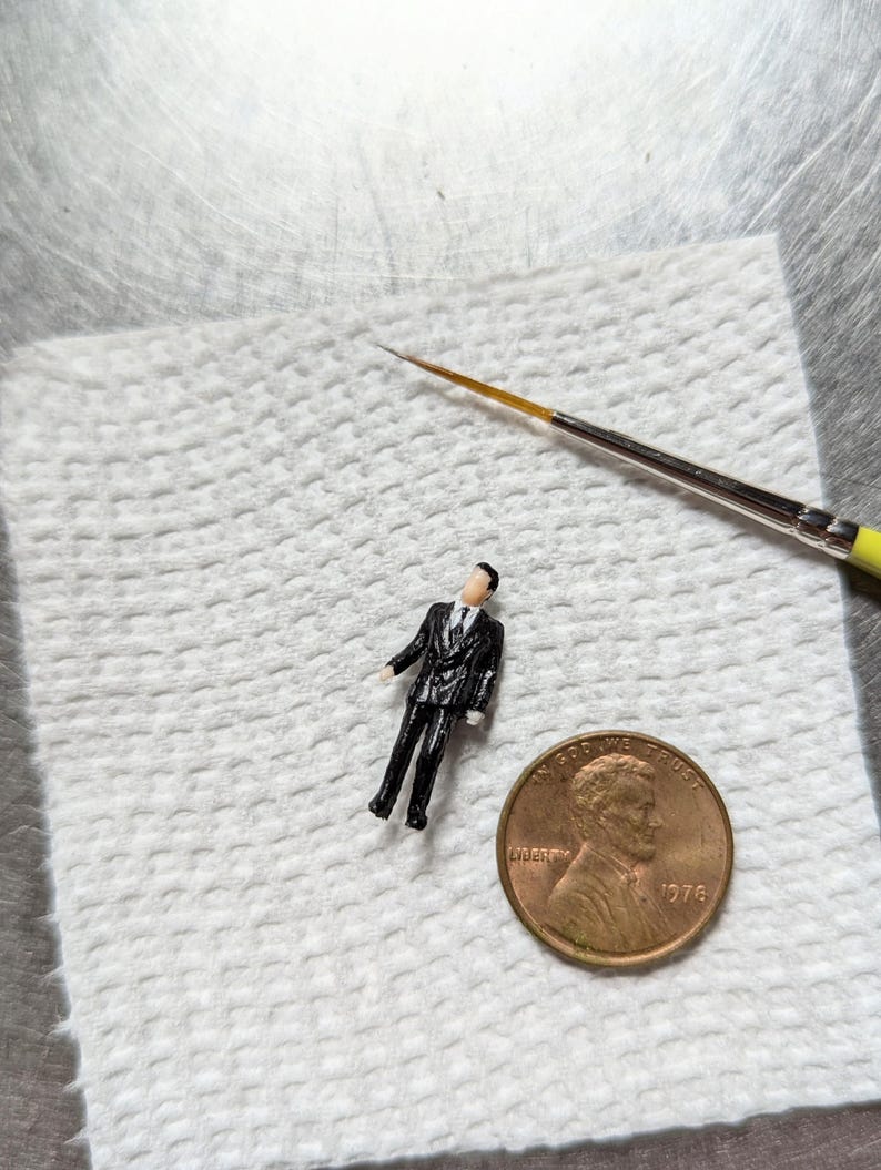 May include: A small, black figurine of a person wearing a suit standing on a white paper towel next to a 1976 US penny.