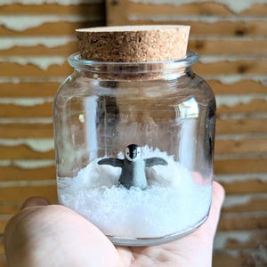 May include: A clear glass jar with a cork stopper containing a small, black and white penguin figurine standing in a bed of white artificial snow. The penguin has its wings spread out to the sides, and the jar is transparent with a smooth surface.