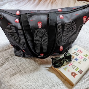 May include: Black tote bag with a white and black graphic print of a person's face. The bag has two black handles and a zipper closure. The bag is sitting on a white blanket with a book and sunglasses on top of it.