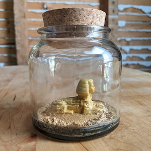 May include: A glass jar with a cork lid containing a gold-colored figurine of a sphinx. The sphinx is sitting on a bed of gold glitter.