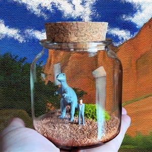 May include: A clear glass jar with a cork stopper containing a miniature blue dinosaur figurine and a small human figure in a suit standing on a bed of brown sand, with a patch of green moss and a white pillar-like object inside the container.