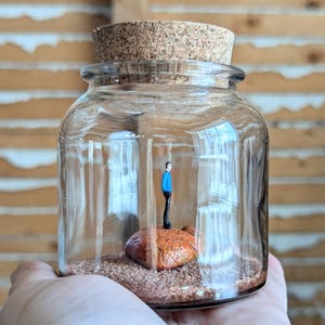 May include: A glass jar with a cork lid contains a miniature figurine of a person standing on a small rock. The jar is filled with brown sand.