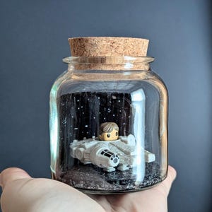 May include: A clear glass jar with a cork stopper containing a miniature white spaceship model and a small figurine with brown hair. The interior features a black background with white speckles resembling stars and a dark textured base.