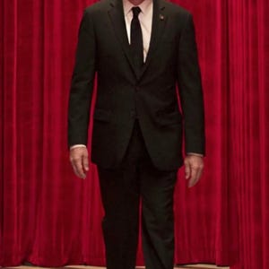 May include: A man in a black suit and tie stands in front of a red curtain with a black and white checkered floor behind him.