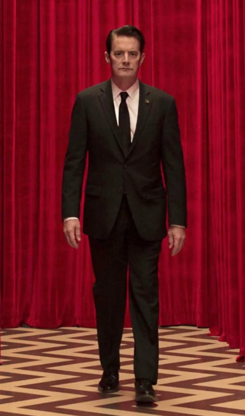 May include: A man in a black suit and tie stands in front of a red curtain with a black and white checkered floor behind him.