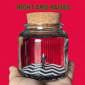 May include: A glass jar with a cork lid containing a miniature scene of a man in a suit standing in front of a red velvet curtain and a black and white checkered floor. The text "RIGHT ARM RAISED" is printed above the jar.