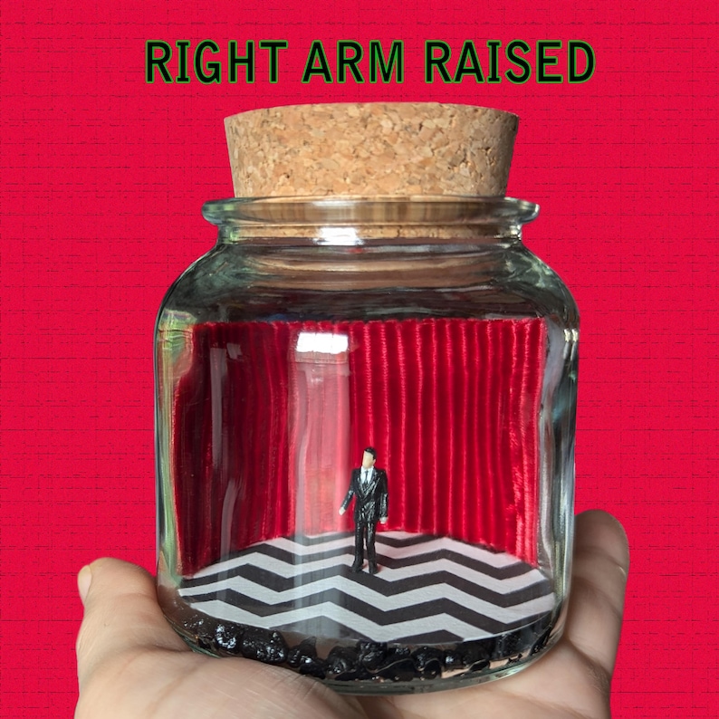 May include: A glass jar with a cork lid containing a miniature scene of a man in a suit standing in front of a red velvet curtain and a black and white checkered floor. The text "RIGHT ARM RAISED" is printed above the jar.
