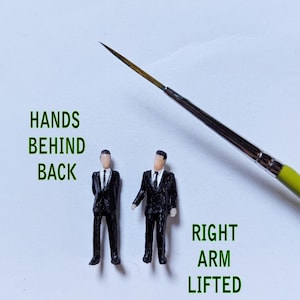 May include: Two miniature figures of men wearing black suits and ties. One figure has their hands behind their back, while the other has their right arm lifted. The figures are standing on a white surface.