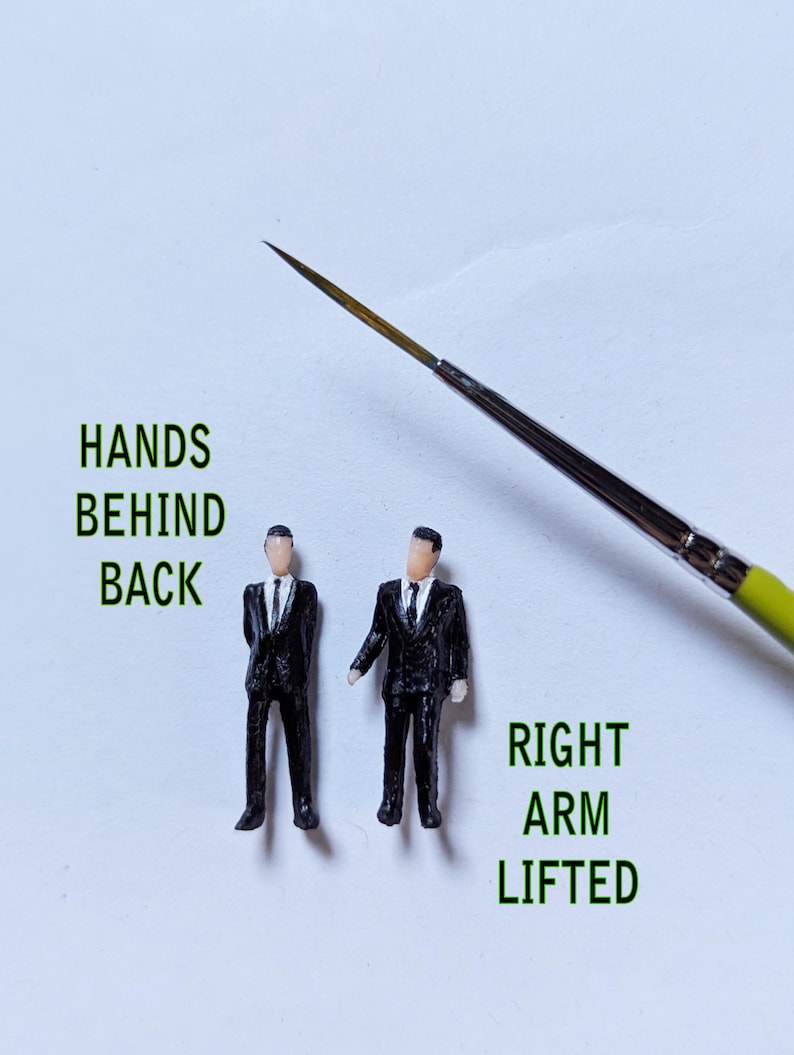 May include: Two miniature figures of men wearing black suits and ties. One figure has their hands behind their back, while the other has their right arm lifted. The figures are standing on a white surface.