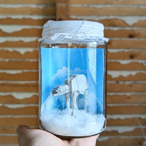 May include: A clear glass jar containing a miniature grey AT-AT walker figure standing on a bed of white artificial snow. The background inside the jar features a blue sky with white clouds, and the lid is covered with white fabric tied with string.