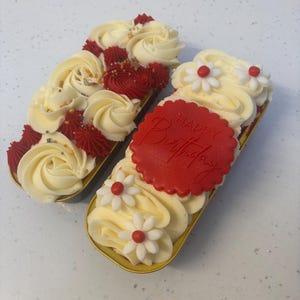 May include: Two rectangular cakes with gold edges, decorated with cream-coloured frosting and red accents. One cake has a "Happy Birthday" message, and the other has red and gold sprinkles. The cakes are decorated with cream-coloured frosting and red accents.