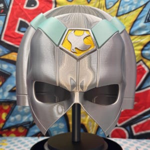 May include: A silver helmet featuring a central pentagonal emblem with a yellow background and a white bird silhouette. The helmet has light blue accents on the upper section and is mounted on a black circular stand with a vertical support rod.