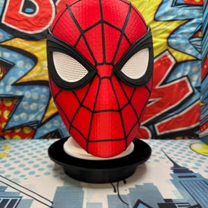 May include: A red and black Spider-Man mask with white eye mesh, displayed on a black stand. The mask features a textured surface and black web detailing. The background includes comic book-style graphics.