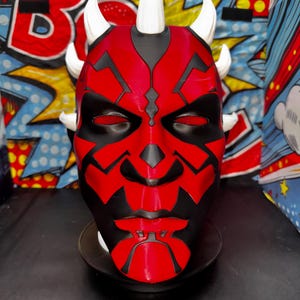 May include: A red and black mask with white horns, resembling a character from a science fiction franchise. The mask features intricate black detailing and red mesh eye coverings. The background includes comic book-style graphics.