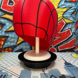 May include: A red Spider-Man mask with black webbing details, displayed on a white stand and a black base. The mask has a textured surface. The background features a comic book-style design.