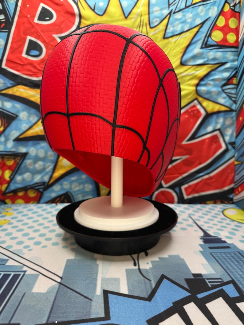 May include: A red Spider-Man mask with black webbing details, displayed on a white stand and a black base. The mask has a textured surface. The background features a comic book-style design.