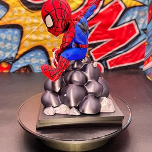 May include: A red, blue, and black Spider-Man figurine perched atop a gray, textured base. The figurine is on a rotating display stand. The background features a colorful comic book-style design.