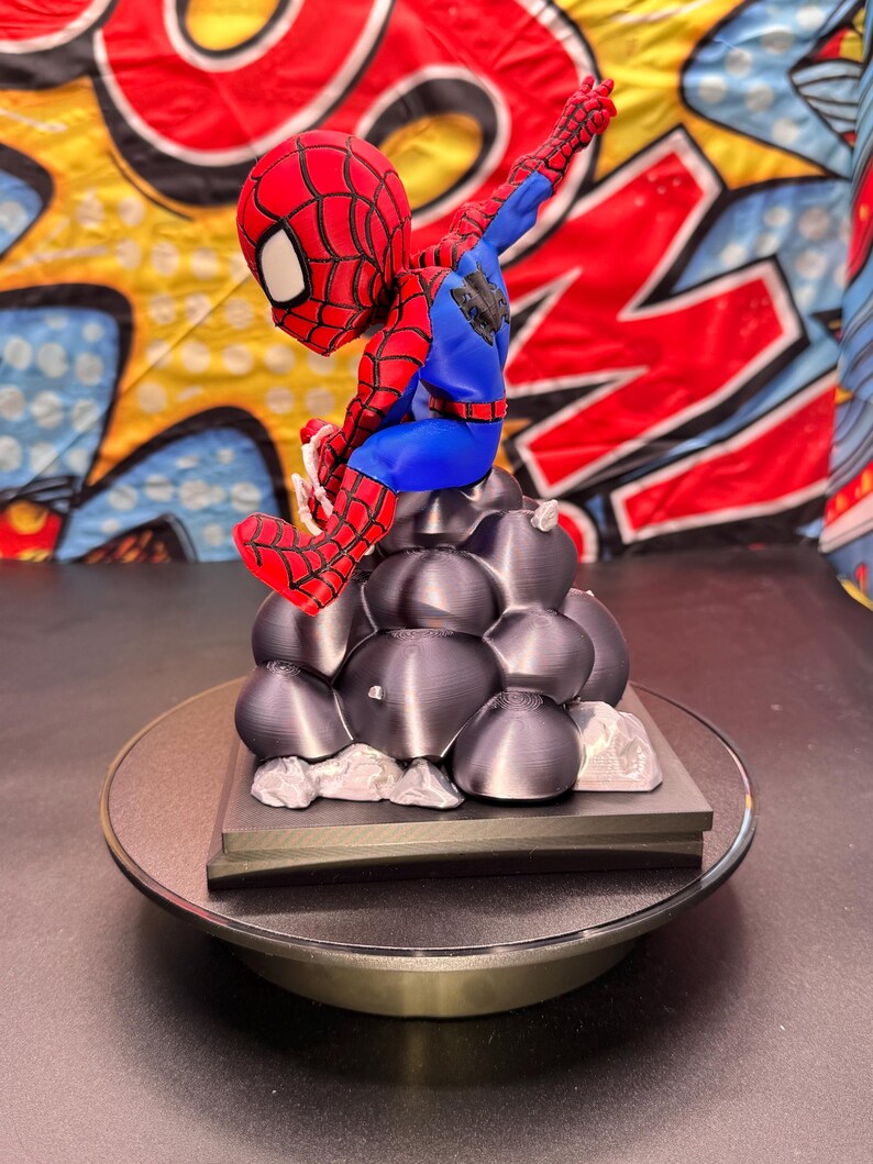 May include: A red, blue, and black Spider-Man figurine perched atop a gray, textured base. The figurine is on a rotating display stand. The background features a colorful comic book-style design.