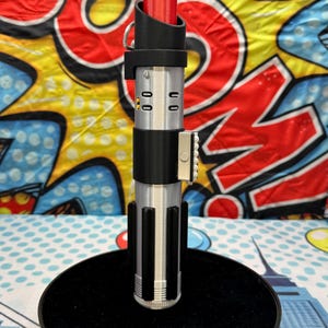 Może przedstawiać: Srebrno-czarny rekwizyt miecza świetlnego z czerwonym ostrzem. Rękojeść ma szczeg&oacute;łowe elementy konstrukcyjne, w tym czarny uchwyt i srebrny emiter. Miecz świetlny jest wyświetlany na czarnym stojaku, na tle kolorowego tła w stylu komiksowym.