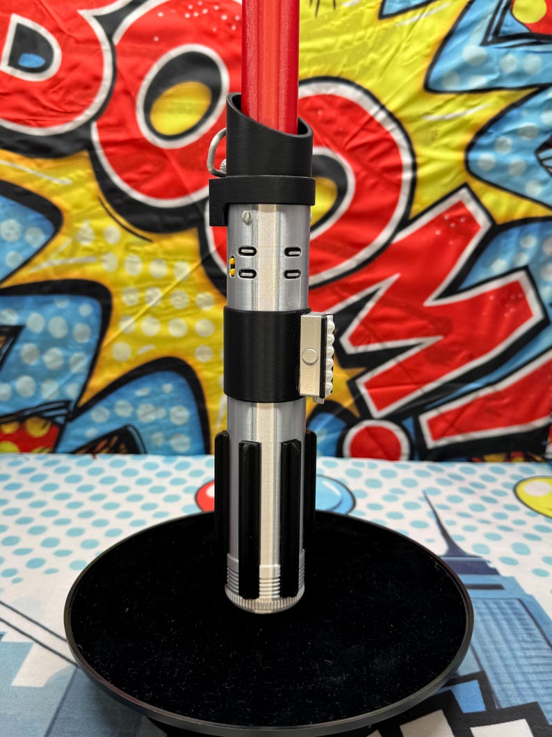 Może przedstawiać: Srebrno-czarny rekwizyt miecza świetlnego z czerwonym ostrzem. Rękojeść ma szczeg&oacute;łowe elementy konstrukcyjne, w tym czarny uchwyt i srebrny emiter. Miecz świetlny jest wyświetlany na czarnym stojaku, na tle kolorowego tła w stylu komiksowym.