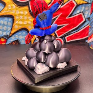 May include: A 3D-printed Spider-Man figurine in a dynamic pose, featuring a red and blue color scheme. The figure is perched atop a cluster of dark gray spheres and a black base, with a rotating display stand.