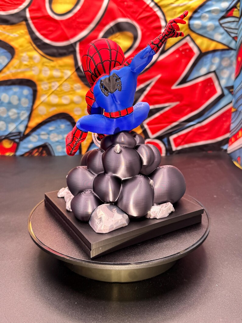 May include: A 3D-printed Spider-Man figurine in a dynamic pose, featuring a red and blue color scheme. The figure is perched atop a cluster of dark gray spheres and a black base, with a rotating display stand.