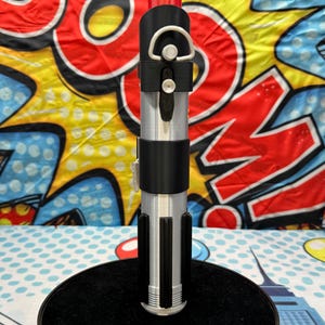 Może przedstawiać: Srebrny i czarny rekwizyt miecza świetlnego, o cylindrycznym kształcie i czarnym uchwycie. Rekwizyt jest prezentowany na czarnym stojaku na tle kolorowego tła pop-art. Miecz świetlny ma srebrny korpus i czarne detale.