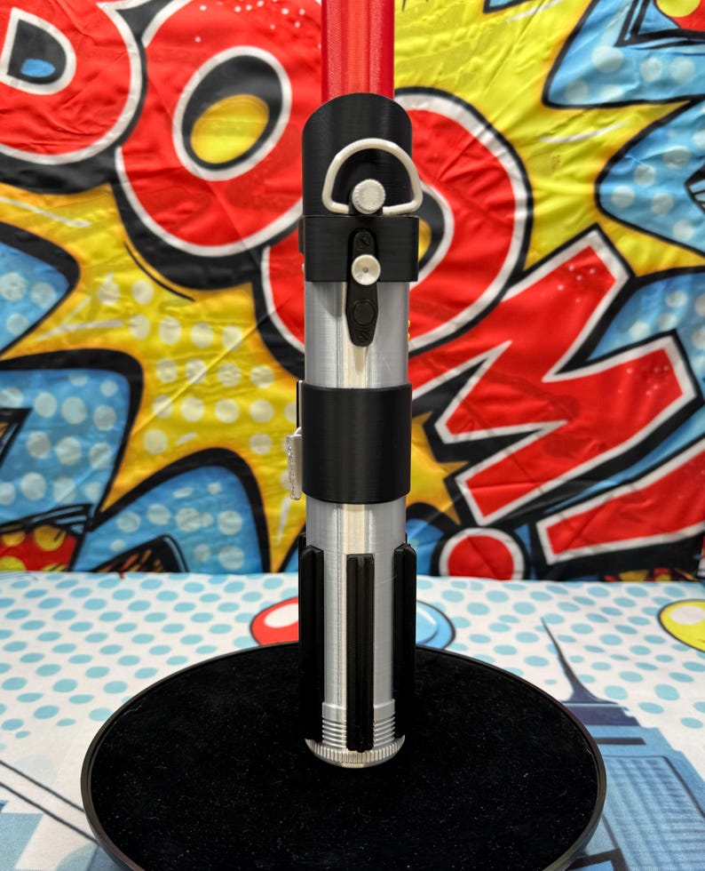 Może przedstawiać: Srebrny i czarny rekwizyt miecza świetlnego, o cylindrycznym kształcie i czarnym uchwycie. Rekwizyt jest prezentowany na czarnym stojaku na tle kolorowego tła pop-art. Miecz świetlny ma srebrny korpus i czarne detale.