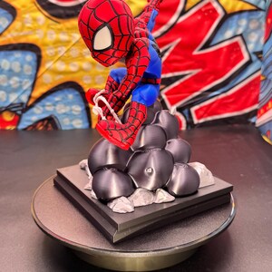 May include: A red, blue, and black Spider-Man figurine in a dynamic pose, perched atop a black base with gray rock-like structures. The figurine is detailed with web patterns and is set against a colorful comic book-style backdrop.