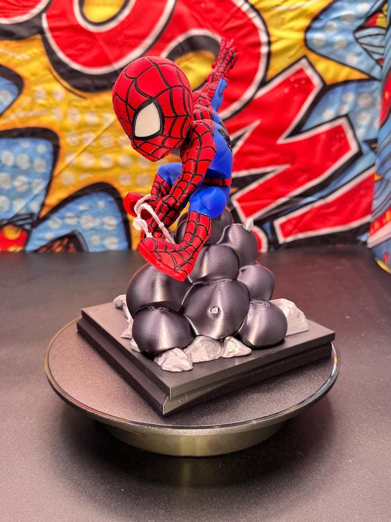 May include: A red, blue, and black Spider-Man figurine in a dynamic pose, perched atop a black base with gray rock-like structures. The figurine is detailed with web patterns and is set against a colorful comic book-style backdrop.