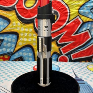 Może przedstawiać: Rekwizyt miecza świetlnego z czerwonym ostrzem i srebrno-czarną rękojeścią. Rękojeść posiada szczeg&oacute;łowe elementy, w tym czarny uchwyt, srebrne akcenty i prostokątny przełącznik aktywacji. Miecz świetlny jest eksponowany na czarnym okrągłym stojaku.