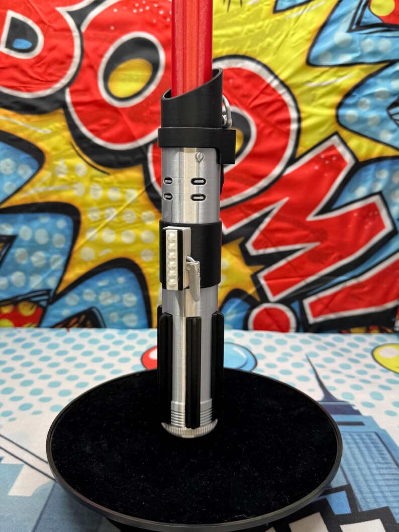 Może przedstawiać: Rekwizyt miecza świetlnego z czerwonym ostrzem i srebrno-czarną rękojeścią. Rękojeść posiada szczeg&oacute;łowe elementy, w tym czarny uchwyt, srebrne akcenty i prostokątny przełącznik aktywacji. Miecz świetlny jest eksponowany na czarnym okrągłym stojaku.