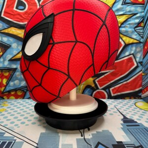 May include: A red Spider-Man mask with black webbing and eye details. The mask is displayed on a white stand with a black base, set against a colorful comic book-style background.