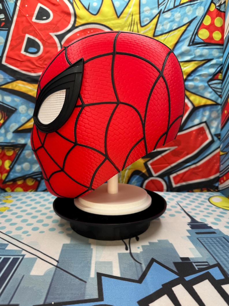 May include: A red Spider-Man mask with black webbing and eye details. The mask is displayed on a white stand with a black base, set against a colorful comic book-style background.