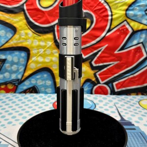 Może przedstawiać: Srebrny i czarny miecz świetlny z czerwonym ostrzem, spoczywający na czarnym, okrągłym stojaku. Miecz świetlny ma szczeg&oacute;łowy uchwyt z pionowymi i poziomymi akcentami. Tło przedstawia kolorowy wz&oacute;r w stylu komiksowym.