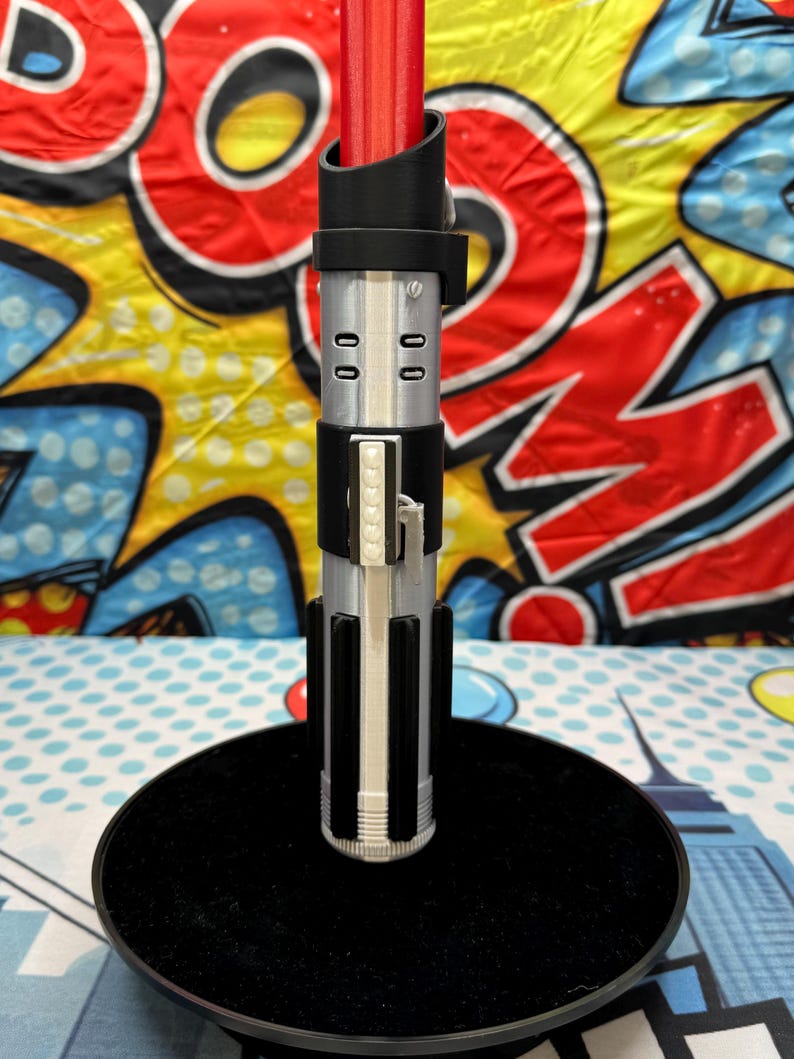 Może przedstawiać: Srebrny i czarny miecz świetlny z czerwonym ostrzem, spoczywający na czarnym, okrągłym stojaku. Miecz świetlny ma szczeg&oacute;łowy uchwyt z pionowymi i poziomymi akcentami. Tło przedstawia kolorowy wz&oacute;r w stylu komiksowym.