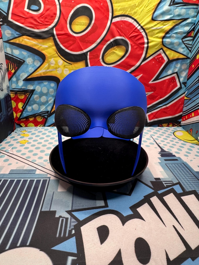 Puede incluir: Una m&aacute;scara de disfraz azul con cubiertas oculares de malla negra y una base negra. La m&aacute;scara est&aacute; sobre una superficie negra. El fondo es un dise&ntilde;o colorido de estilo c&oacute;mic.