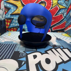 Puede incluir: Una m&aacute;scara de superh&eacute;roe azul con grandes aberturas para los ojos cubiertas de malla. La m&aacute;scara se exhibe sobre un soporte negro contra un fondo de gr&aacute;ficos de estilo c&oacute;mic. La m&aacute;scara tiene un acabado liso y est&aacute; dise&ntilde;ada para cubrir la parte superior de la cara.